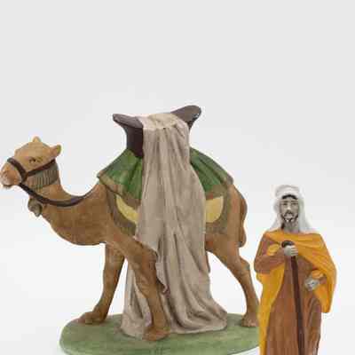 One set of the camel and male Bedouin figurine.: Image of one of the male figurines wearing a brown traditional Kuwaiti dresses, a yellow shall, and white head covering with a walking stick. The camel has a yellow and green saddle pad, a large brown saddle, and draped beige cloth. The reins are brown. The male figurine is standing in front of a the camel.