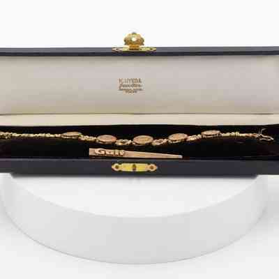 Box for tie bar and bracelet: A long, velvet-lined black jewelry case. Inside it has a matching bracelet with five charms lying next to the tie bar. The lid of the case has the jewelers name.