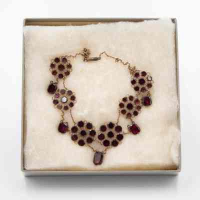 Gold and garnet necklace with flower-shaped design, in box.: The image showcases a gold necklace with garnet stones arranged in a flower-shaped design, resting in a soft, cushioned box.