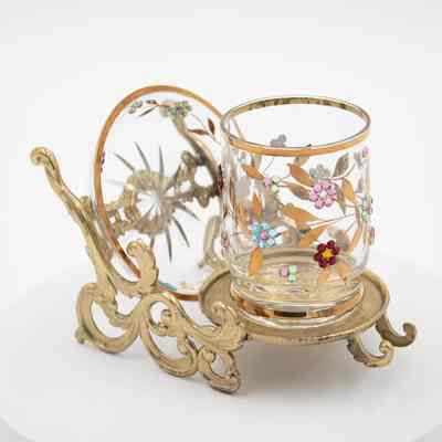 Glass cup and dish set on gold sleigh from Japan.: Up close image of a glass cup sitting on a gold sleigh with glass saucer on the back. The cup and saucer are decorated with gemstone flowers and gold leaves. The sleigh has gold swirls.
