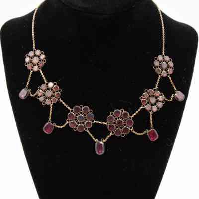 Gold and garnet necklace with flower-shaped design, on stand.: The necklace features intricate flower-shaped designs crafted from gold and garnet, displayed on a black stand.