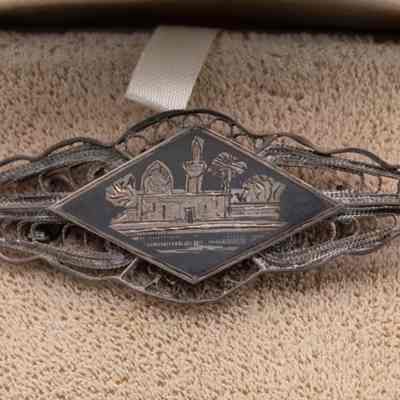 Diamond-shaped silver pin with oasis scene: A diamond-shaped silver pin intricately displays an engraved oasis scene with palm trees and structures, set against a textured background.