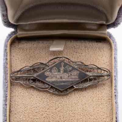 Diamond-shaped silver pin with oasis scene: A diamond-shaped silver pin intricately displays an engraved oasis scene with palm trees and structures, set against a textured background.