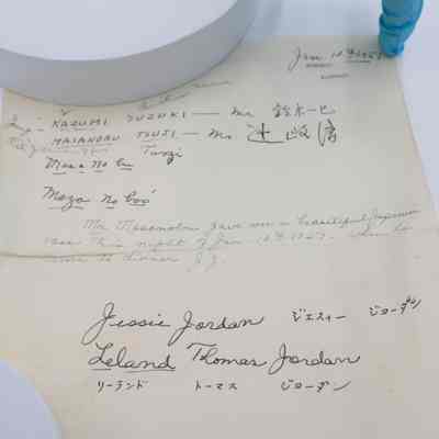 Note associated with the Japanese cloisonne floral vase given to the Jordans by Masanobu Tsuji.: Up close image of the handwritten note on yellowing paper with black handwriting. The top left corner is dated "Jan. 10, 1957" and "Ahmadi, Kuwait" is listed as the location. There are also two names listed in both English and Japanese: "Kazumi Suzuki" and "Masanobu Tsuji." There is then a note written by Mrs. Jessie Jordan that reads "Mr. Masanobu gave me a beautiful Japanese vase this night of Jan. 10 1957 - when he came to dinner. J.J." At the bottom of the page, there are the Jordans' names written in both English and Japanese, "Jessie Jordan" and "Leland Thomas Jordan."