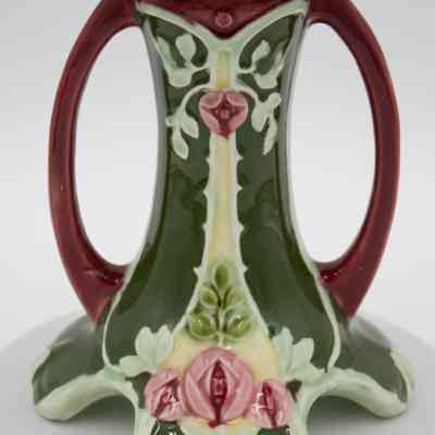 Pedestal base of the green ceramic vase with pink flowers.: Up close image of the bottom of the pedestal vase. There are three pink flowers, likely roses, in the center. There are also leaves in two shades of green.
