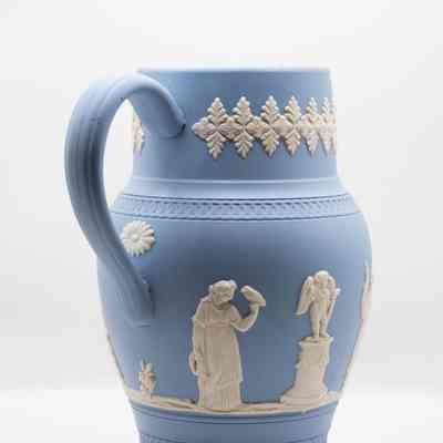 Wedgwood Jasperware pitcher: The Wedgwood blue Jasperware pitcher features intricate white relief designs, including a small flower under the handle. This side of the pitcher features a woman holding a harp and a bird, as well as a cherub on a pedestal.