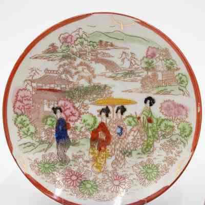Hand-painted Japanese porcelain saucer.: Up close image of the painted eggshell porcelain saucer. It depicts a group of geisha in a traditional Japanese garden. The geisha are dressed in red, yellow, blue, and orange and are surrounded by pink and green flowers/trees. The saucer has a red rim and handle.