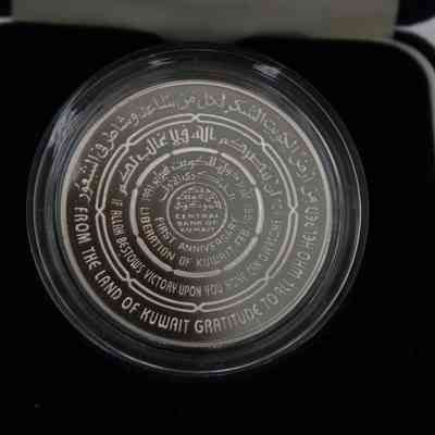 Commemorative Coin: Close-up image of the commemorative coin. The coin is sliver and is in a clear plastic case. The coin is encased in black felt. In capital white letters expanding out from the middle and occupying the bottom half, the coin reads: "Central Bank of Kuwait | First Anniversary Liberation of Kuwait Feb. 1991 | If Allah Bestows Victory Upon You None Can Overcome You | From The Land of Kuwait Gratitude to All Who Helped." The top half of the coin has the same inscription but in Arabic.