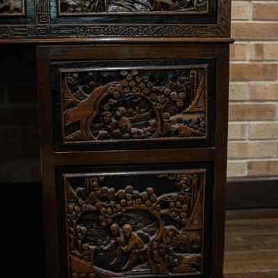 Decorative panels on wooden desk: The image displays a section of a carved wooden desk from Hong Kong, showcasing intricate, detailed carvings of people and landscapes on the drawer fronts, set against a brick wall.
