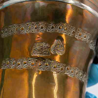 Text on coffee pot: A copper coffee pot with leather-wrapped handle, brass details, and stamped Arabic script.