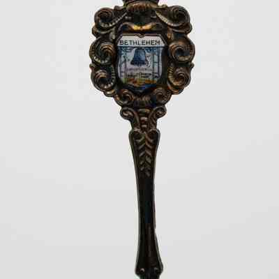 One of the collector's spoons from Bethlehem.: Image of the silver collector's spoon decorated with the Christmas Bell on the handle. The background is white enamel with a townscape and bell in the center, the top of the design reads "BETHLEHEM" while the center reads "CHRISTMAS BELL." There is also silver details around the shield and handle of the spoon in swirls designs.