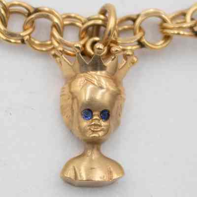 Charm of the bust of a boy with a crown and blue gem eyes on Mrs. Jessie Jordan's charm bracelet.: Up close image of the gold charm of a bust of a boy with a pointed circular crown and blue gem stone eyes.