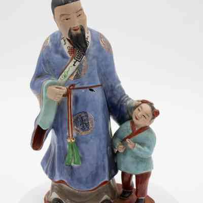 Painted porcelain figurine of Mongolian man and boy.: Image of a porcelain figurine of a Mongolian man dressed in a blue robe with gold foil Mongolian symbols on it. He stands above a little boy who is wearing a light blue top with red details and red pants. The man's hair is in a bun atop his head with a blue covering and the boy has two buns both with red coverings. They are standing on a red base.
