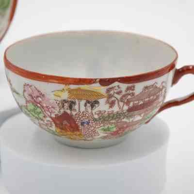 Hand-painted Japanese porcelain teacup.: Up close image of the painted eggshell porcelain teacup. It depicts a group of geisha in a traditional Japanese garden. The geisha are dressed in red, yellow, and orange and are surrounded by pink and green flowers/trees. The saucer has a red rim and handle.