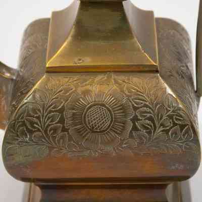 Base of coffee pot: The image depicts the base of a large brass coffee pot, intricately engraved with a detailed floral design, featuring leaves and a prominent central flower.