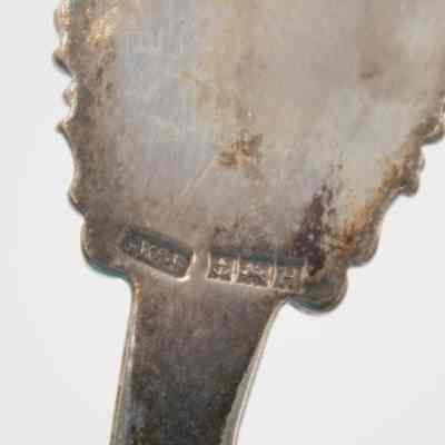 Back of the souvenir spoon with silversmith marks.: Up close image of the back of the silver souvenir spoon. The silversmith marks indicate it was made by George, Kenning & Son, The Original British Manufactures of the Highest Quality Masonic Regalia and Accessories in Birmingham, England. From left to right, the stamped symbols are "GK&S" indicating the company, an anchor indicating the city of Birmingham, a lion indicating it was made in England, and a "H" indicating the year it was produced.