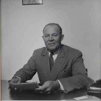 Photo of Mr. L.T. Jordan sitting at a desk holding papers and a pen.: Black and white image of Mr. L.T. Jordan sitting at a desk against a light wall holding papers and a pen. The frame is yellow.