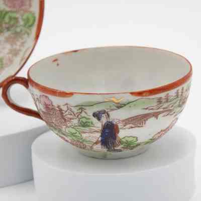 Hand-painted Japanese porcelain teacup.: Up close image of the painted eggshell porcelain teacup. It depicts a group of geisha in a traditional Japanese garden. The geisha is dressed in blue and is surrounded by pink and green flowers/trees. The saucer has a red rim and handle.