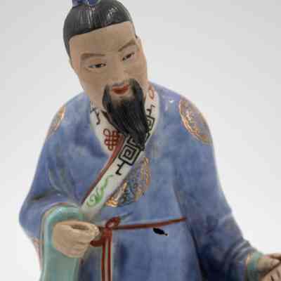 Painted porcelain figurine of a Mongolian man.: Up close image of the porcelain Mongolian man dressed in a blue robe with gold details. The man has a black beard and black hair pulled into a bun with a matching blue covering.
