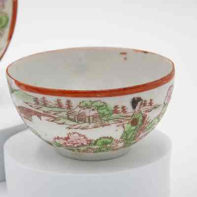 Hand-painted Japanese porcelain teacup.: Up close image of the painted eggshell porcelain teacup. It depicts a group of geisha in a traditional Japanese garden. The geisha is dressed in green and is surrounded by pink and green flowers/trees. The saucer has a red rim and handle.