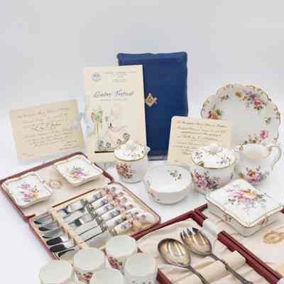 Complete collection of Royal Crown Derby porcelain given to Mrs. Jessie Jordan from Masonic Lodges.: Image of the the full set of Royal Crown Derby items received by Mrs. Jessie Jordan. There is a blue Masonic Presentation Bible propped up in the back along with three invitations from various Ladies' Nights. All of the porcelain items are white and have the same floral pattern decorating them. There is one larger bowl, a cream pitcher, two sugar dishes (one large, one small), a smaller bowl, two cigarette ashtrays, a cigarette box, a set of six silver knives in a red case, a pair of silver salad serving spoons in a red case, and six small teacups.