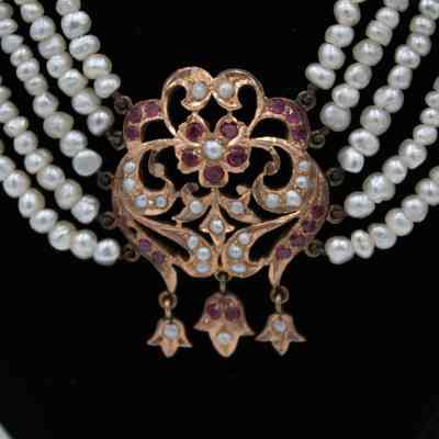 Gold detail at the center of the necklace. It is embedded with red gemstones and pearls and has three drop pendants.: Up close image of the gold center pendant of the pearl necklace. Five pearl strands hold up each side of the pendant. There is a gold flower in the center with leaves and swirls around. There are embedded red gemstones and pearls in the pendant. There are three small connected pendants dangling from the bottom in a leaf shape.