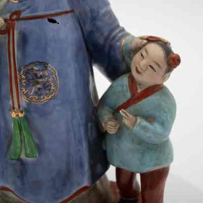 Painted porcelain figurine of a Mongolian boy.: Up close image of the porcelain Mongolian boy dressed in a light blue shirt with red details and matching red pants. The boy has black hair pulled into two buns with red coverings. He is looking up at the man admiringly.