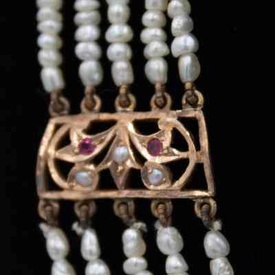 Gold detail on the gold and pearl necklace.: Up close image of one of the gold details on the 5-strand necklace. The gold charm has two red stones and three pearls embedded in it.