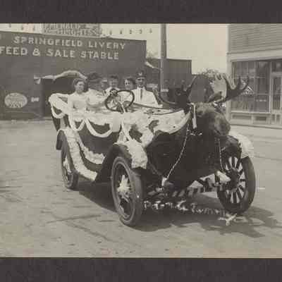 Photograph of old car decorated with a moose head and bunting, possible a parade float, with two men in front and two women and a boy in the back seat. Out in front of Springfield Livery Feed & Sale Stable. Date unknown.