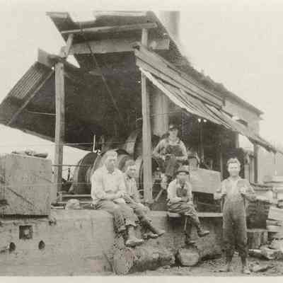 Photograph of five men crew seated and standing in front of a covered steam donkey