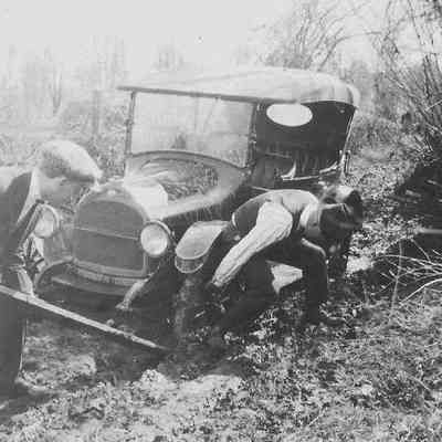 Photograph of two men trying to lever car out of mud with a long board. Date unknown.