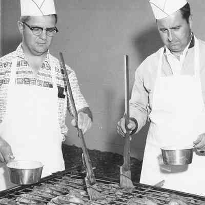 Photograph of two unknown men basting chicken, date unknown.
