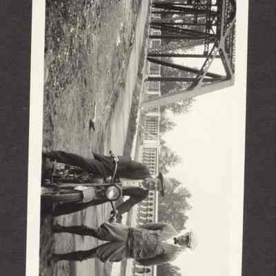 Photograph of two men one standing and one sitting on a motorcycle with bridge in background. Date unknown.