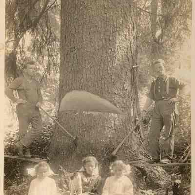 Photograph of two lumberjacks standing with three girls sitting at the base of a tree with their dog