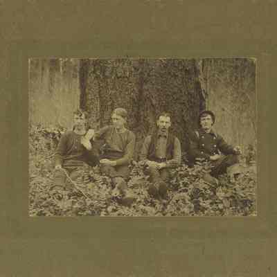 Photograph of four loggers sitting at base of tree, circa 1890-1910