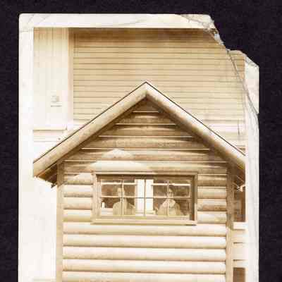 Photograph of small house with people cutouts inside, outside an unknown lumber company entrance, date unknown