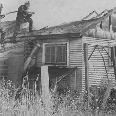 Photograph of firemen spraying roof of small house on fire, date unknown.