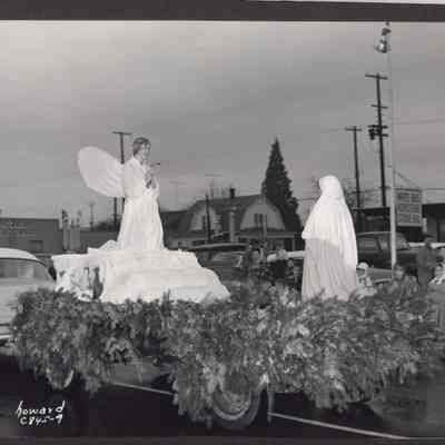 Photograph of flat bed trailer with pine boughs, parade float with two figures. Date unknown.