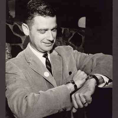 Photograph of unknown man in a suit with two watches, one on each wrist, looking at them. Date unknown.