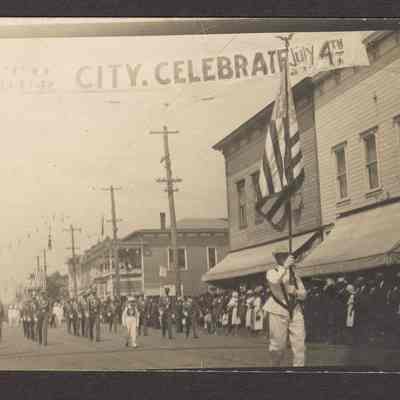 Photograph of a Postcard of a parade with banner over the street: "Come to our City. Celebrate July 4th." About 1916, looking west toward 3nd and Main St. Opera House and Springfield Hotel with Brownsville railroad between at right corners.