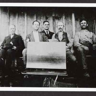Photograph of five men sitting behind a chalk board