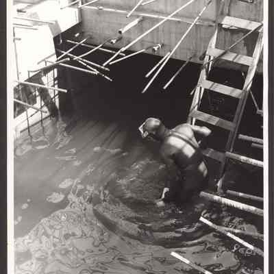 Photograph of unknown man in a wet suit looking into water at bridge construction worksite. Date unknown.