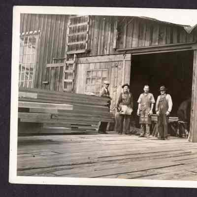Photograph of four unknown men standing in lumber yard outside of a building, date unknown