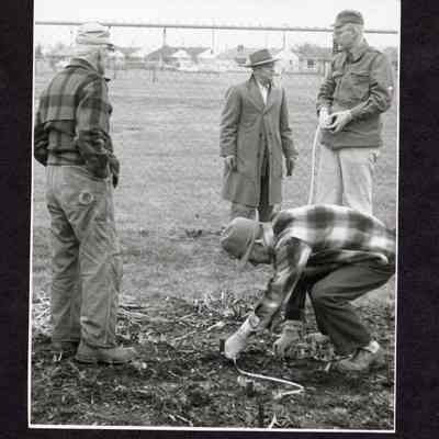 Photograph, 4 men measuring ground in front of chain link fence, date and location unknown.