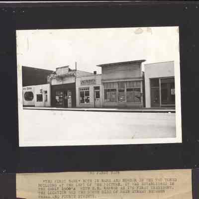 Photograph of the First Bank. "The First Bank" both in name and number is the two-toned building at left of the picture. It was established in the early 1900s with E.E. Kepner as its first president. The location was south side of Main St. between Third and Fourth St.