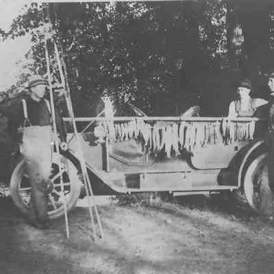 Photograph of Jess Seavey and two men with an early car and a pole of fish, date unknown