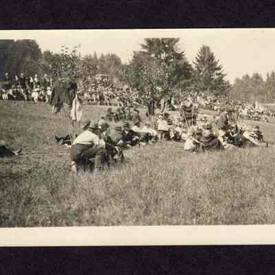 Photograph on postcard of people sitting, talking and looking at something at the lodge picnic at Camp Creek School