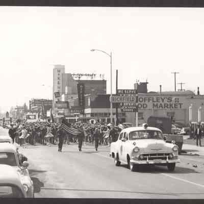 Photograph of a parade lead by 1950s police car, Boy Scouts with colors, and a marching band. 7th and Main Looking West. Date unknown.