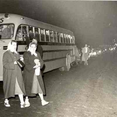 Photograph of two students walking in front of a bus, date unknown.