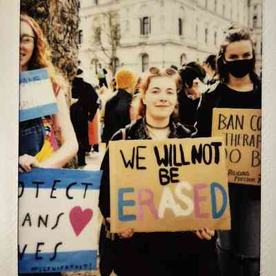 A group of people hold signs at a protest, with supportive messages such as "WE WILL NOT BE ERASED".